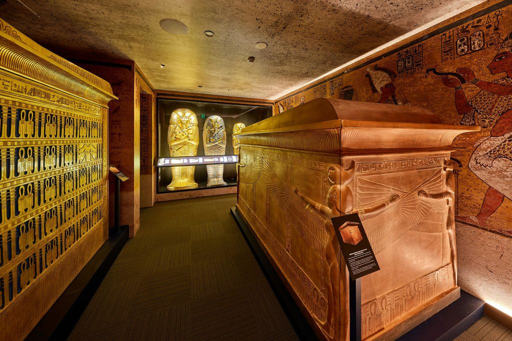 이집트 석관, 상형문자가 새겨진 벽, 조명이 켜진 조각상 두 개를 배경으로 한 박물관 전시관입니다.
