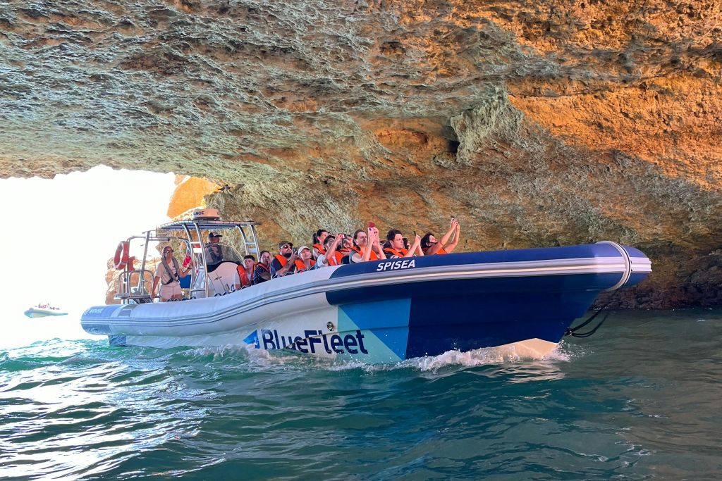 블루플리트 보트에 주황색 구명조끼를 입고 물 위 바위 벽이 있는 동굴을 탐험하는 사람들입니다.