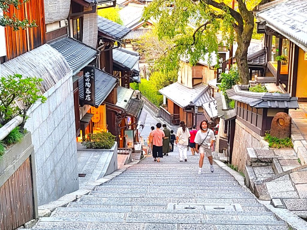 일본 전통 건물, 보행자 몇 명이 걷는 비탈진 자갈길, 카메라를 보며 미소를 짓는 사람.