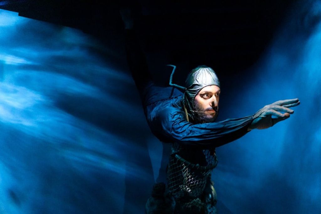 금속 의상과 헤드기어를 입은 사람이 수중 장면을 닮은 파란색 조명 아래에서 극적인 포즈를 취하고 있습니다.