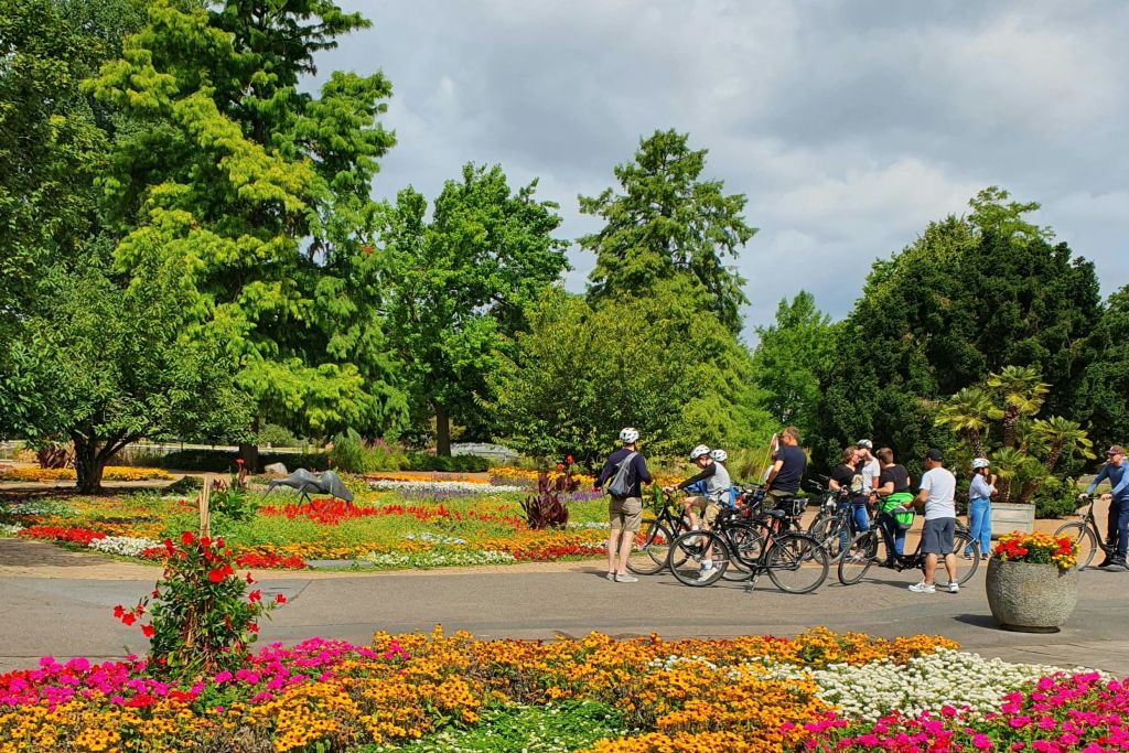 활기찬 화단과 높은 녹색 나무가 있는 다채로운 정원의 길에 자전거를 탄 사람들이 모여듭니다.