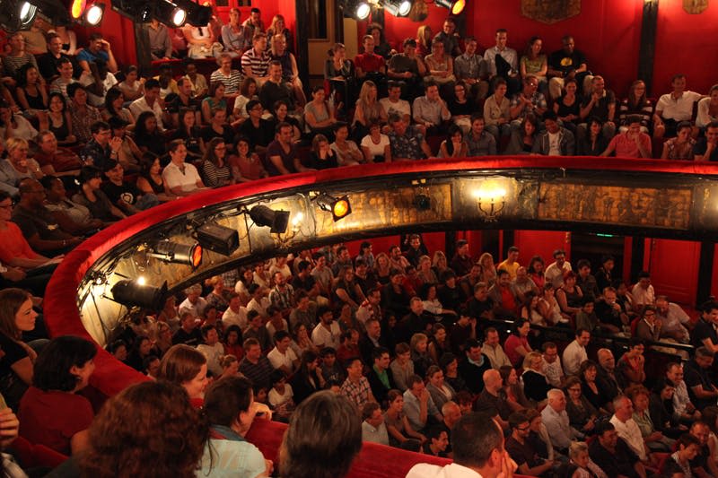 여러 무대 조명이 켜진 빨간색 좌석이 있는 원형 극장에는 많은 관객이 앉아 보이지 않는 공연에 집중하고 있습니다.