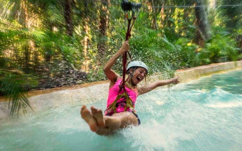 분홍색 셔츠와 헬멧을 착용한 여성이 초록색 숲을 배경으로 물에 기쁘게 지퍼로 잠그고 있습니다.