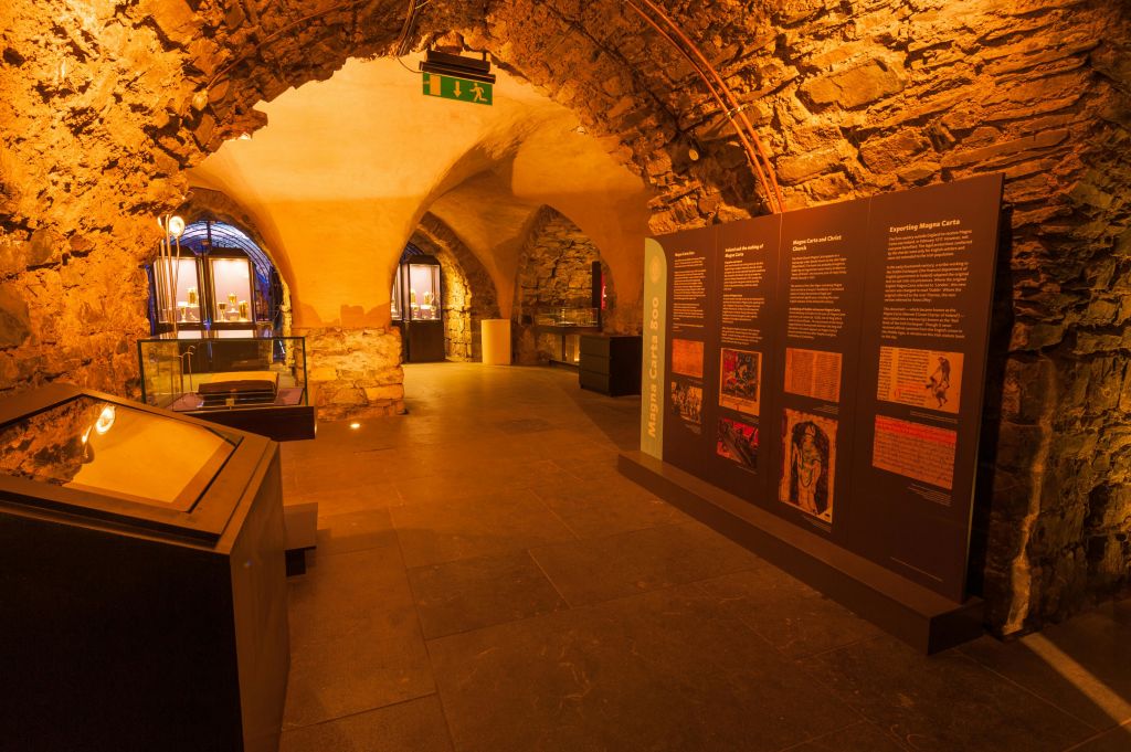 안내 패널, 아치형 통로, 유리 진열대, 녹색 출구 표지판이 있는 석조 박물관 전시실입니다.