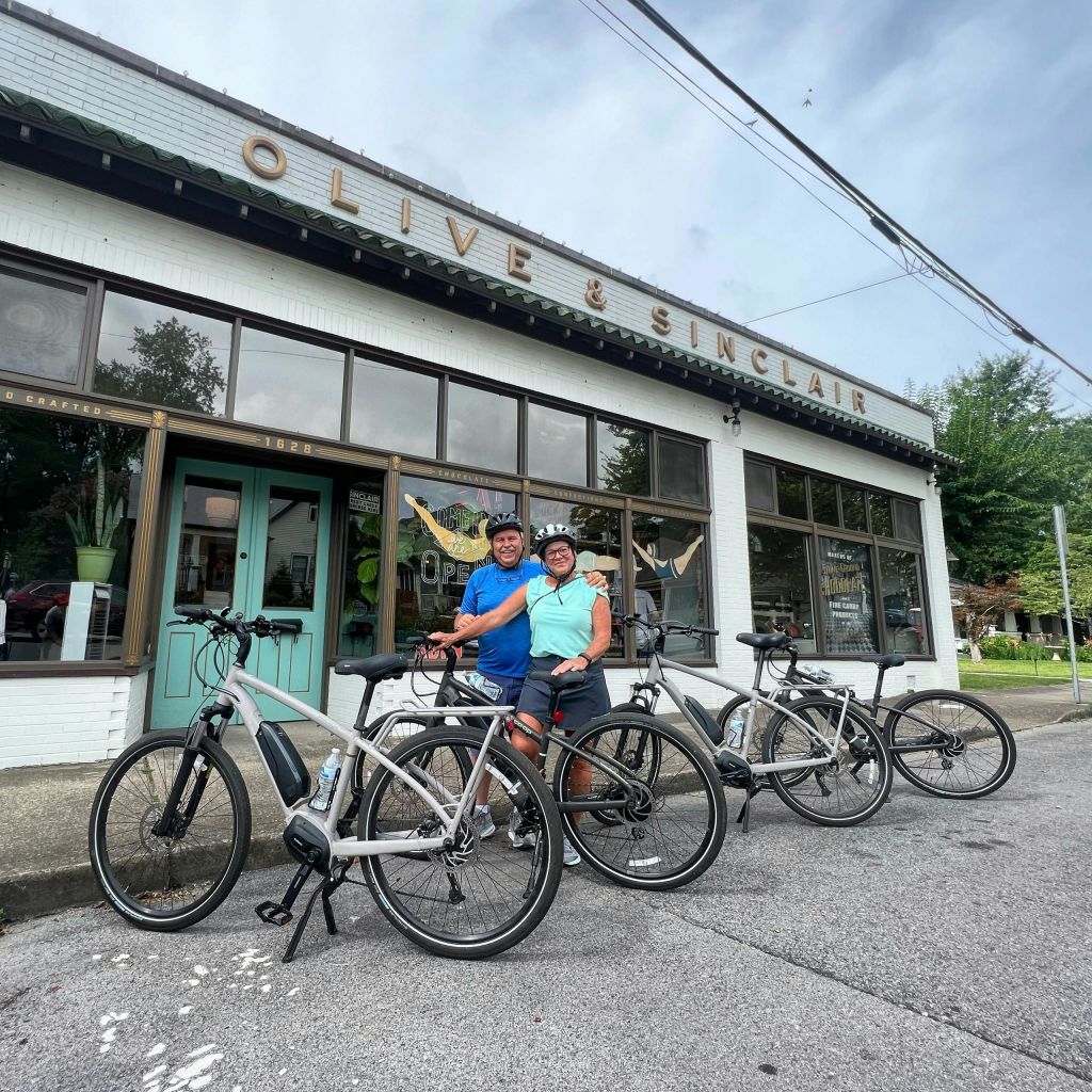 "Olive & Sinclair" 표지판이 있는 건물 밖에 자전거를 탄 두 사람이 서 있습니다. 이 장면에는 거리와 녹지가 포함되어 있습니다.