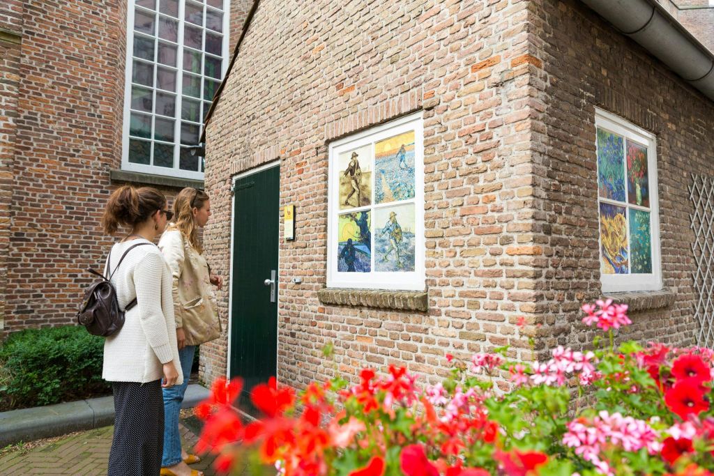 두 여성이 초록색 문 옆 벽돌 벽에 걸린 다채로운 그림 세트를 바라보고 있는데, 빨간색 꽃이 앞쪽에 피어 있습니다.