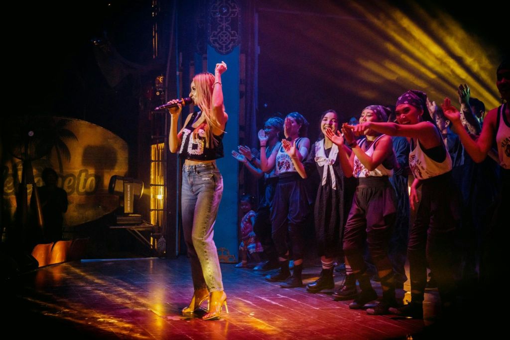 무대에서 한 여성이 조율된 의상을 입고 박수를 치며 환호하는 공연자들의 무리에 둘러싸여 마이크를 잡으며 노래하고 있습니다.