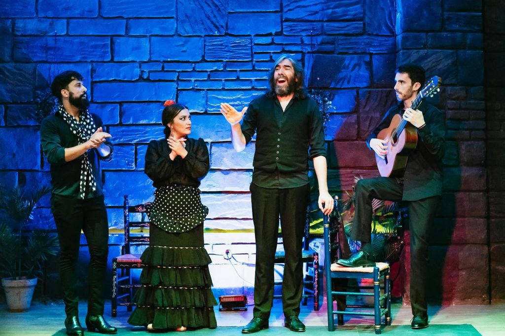 세 명의 연주자가 검은색 옷을 입고, 한 명은 노래를 부르고, 한 명은 기타를 치고, 한 명은 박수를 치고 있습니다. 다채로운 조명이 있는 돌 벽 배경.