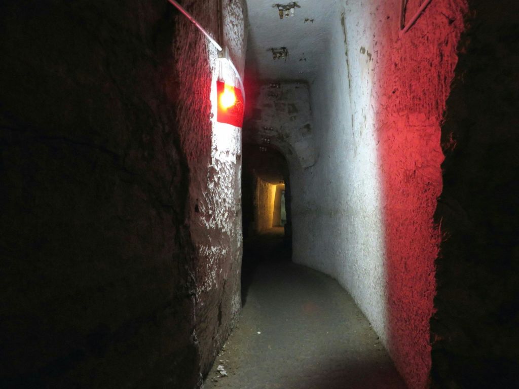 거친 벽, 좁은 통로, 벽에 빨간색 조명기구가 있는 조명이 희미한 지하 터널입니다.