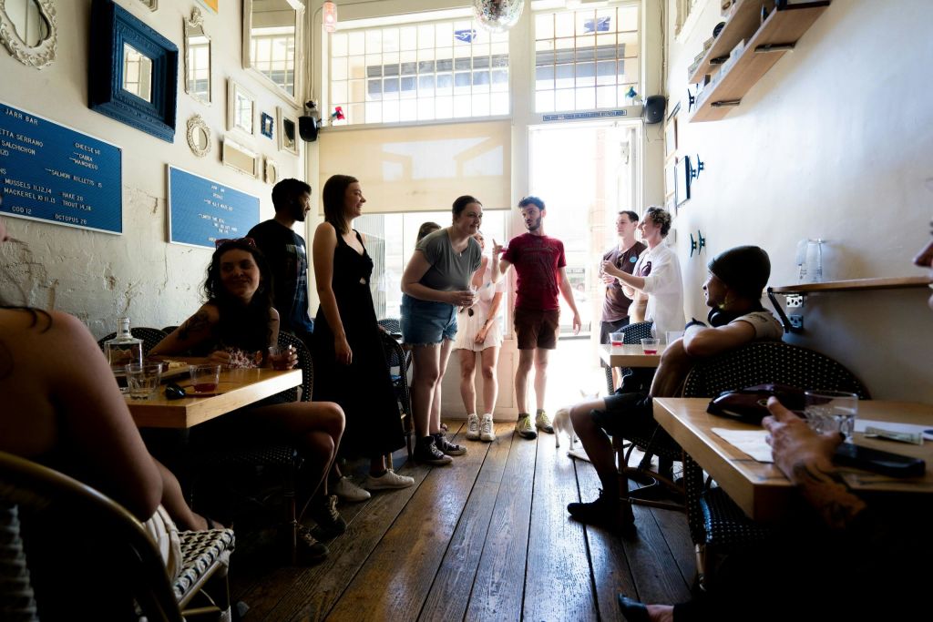 나무 바닥과 벽 선반이 있는 햇살이 내리쬐는 카페에서 사람들과 어울리며 입구 근처에 앉아서 서 있습니다.