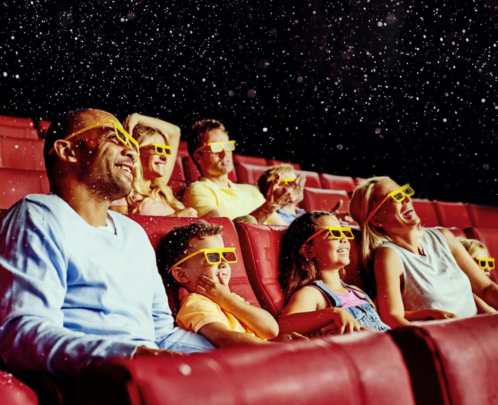 3D 안경을 쓰고 빨간 극장 좌석에 앉아 미소를 지으며 공중에 떠 있는 입자를 올려다보는 사람들.