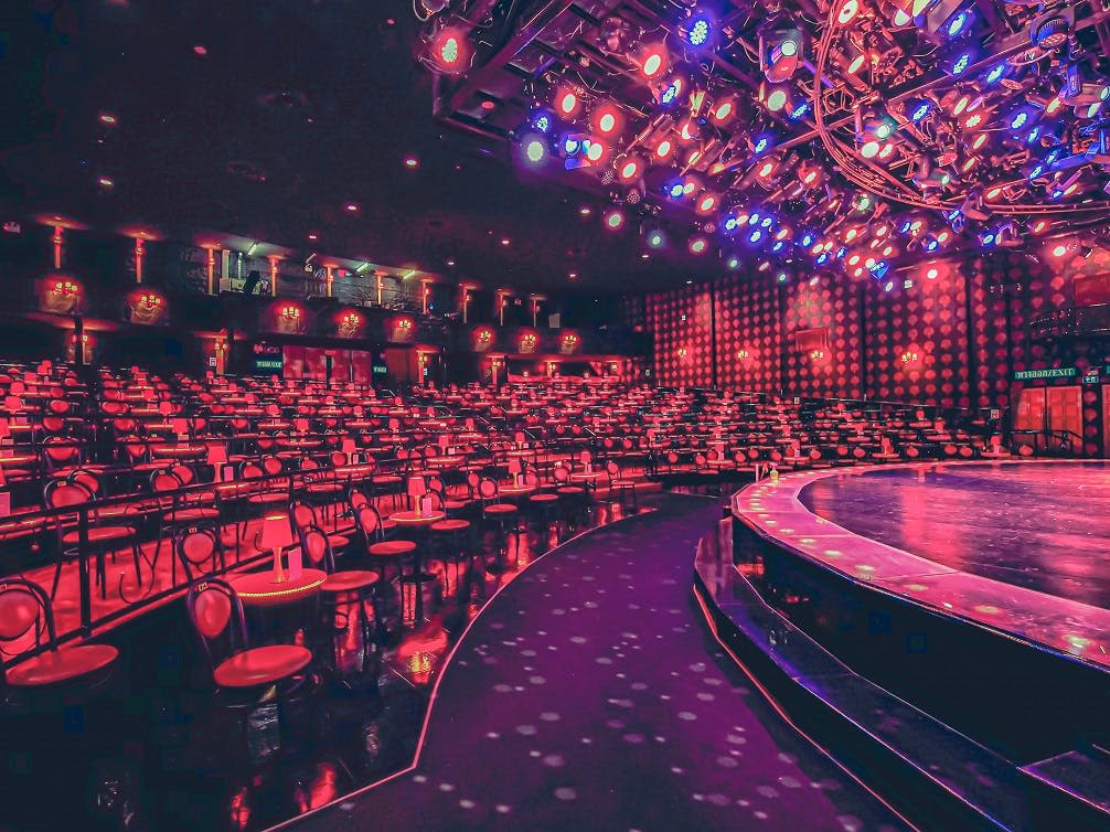 화려한 무대 조명 아래 원형 무대를 향하여 원형 테이블과 빨간색 의자가 줄지어 놓인 활기차고 텅 빈 극장.