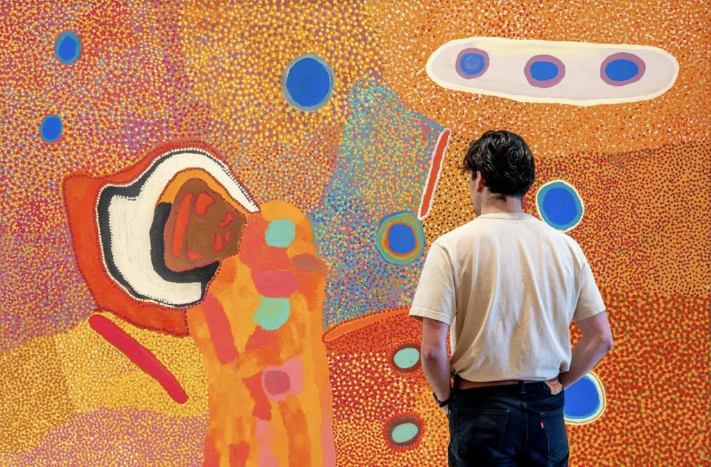 흰색 셔츠를 입은 사람이 따뜻한 톤의 점, 원, 유기적인 모양이 특징인 다채로운 추상화를 관찰합니다.