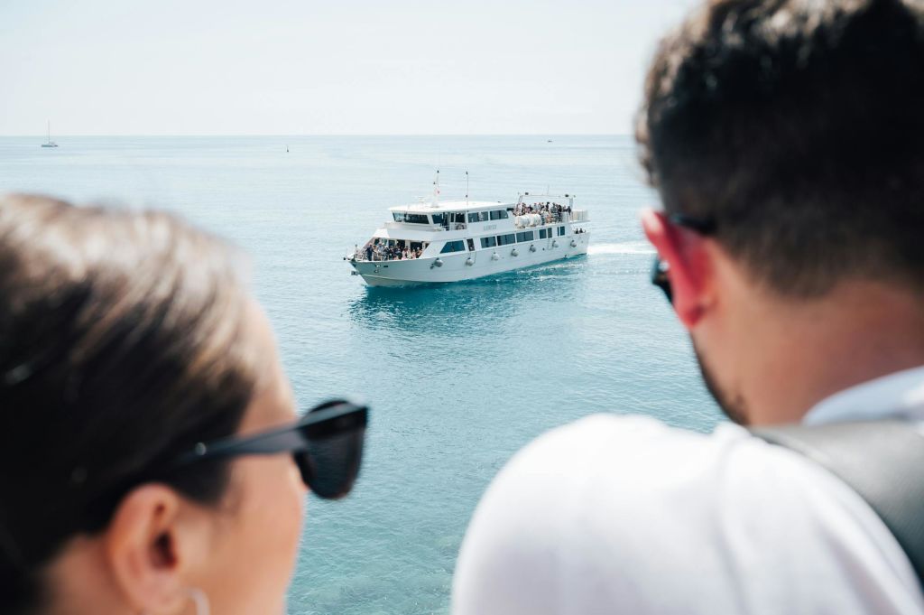 쌍안경을 든 남성이 잔잔한 바다를 항해하는 승객들로 가득 찬 흰색 페리 보트를 보고 있습니다.