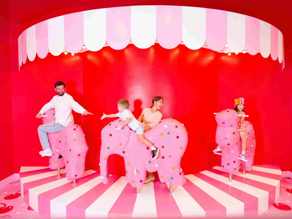 회전목마의 분홍색, 물방울무늬, 코끼리 모양의 좌석에 앉아 있는 세 사람은 밝은 조명, 빨간색 및 분홍색 카니발 세팅을 하고 있습니다.
