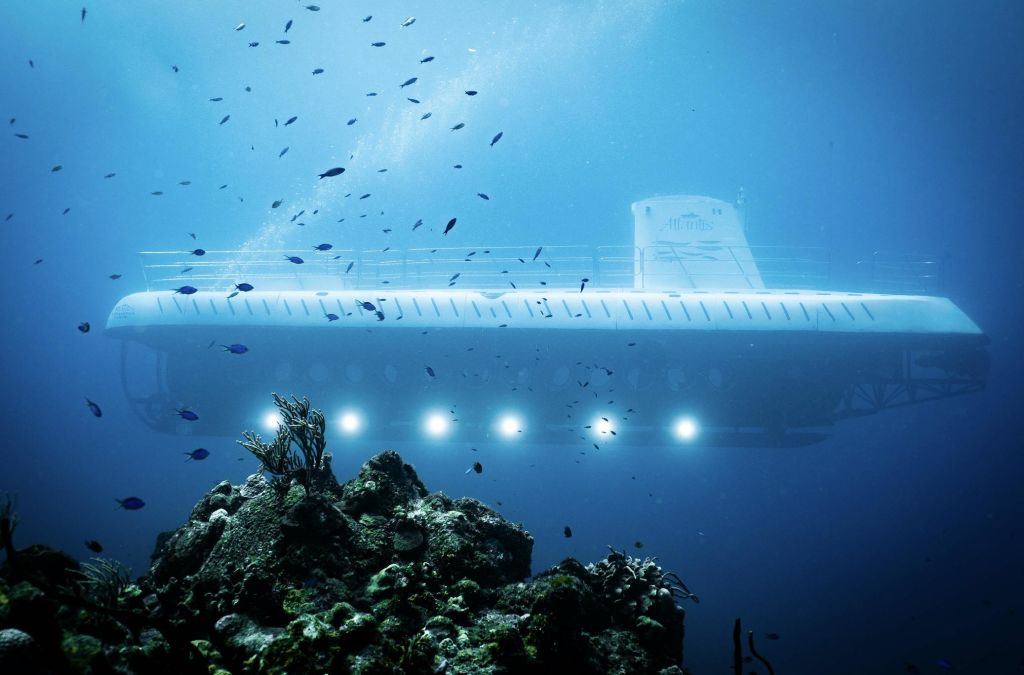 반짝이는 창문이 있는 물에 잠긴 하얀 잠수함이 바위산호를 지나 맑고 푸른 물에 둘러싸인 작은 물고기들로 둘러싸여 있습니다.