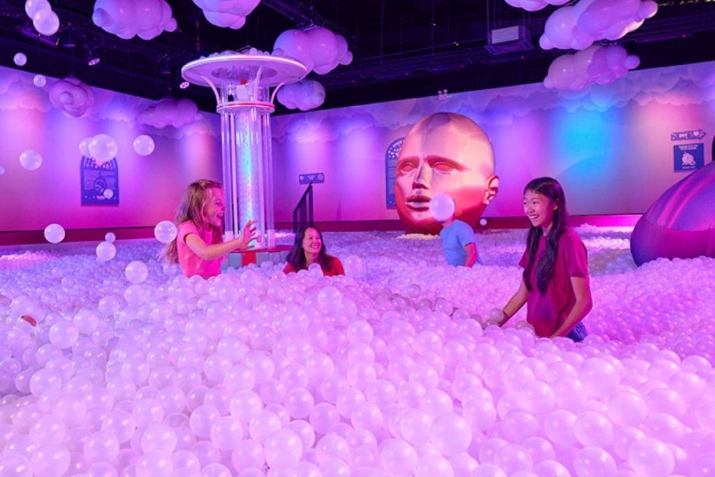 밝은 흰색 공, 화려한 벽, 커다란 머리 조각을 배경으로 커다란 공 구덩이에 서 있는 세 사람.