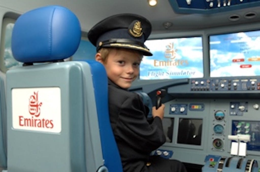 파일럿 유니폼을 입은 어린이가 비행 시뮬레이터 조종석에 앉아 웃으며 컨트롤 스틱을 들고 있습니다. "에미레이트 항공 비행 시뮬레이터" 로고가 표시됩니다.