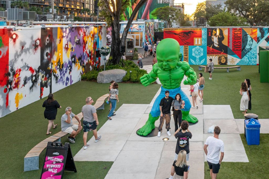 화려한 그라피티 벽을 배경으로 공원의 커다란 녹색 헐크 동상 근처에서 걷고 포즈를 취하는 사람들.