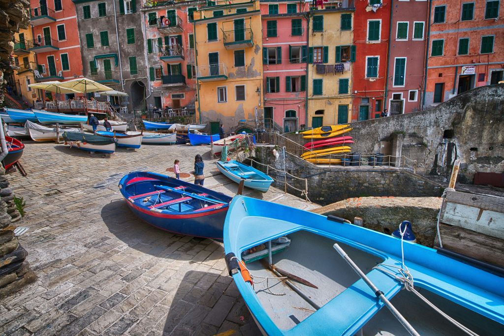 돌 부두에 다채로운 보트가 주차되어 있으며 근처를 걷는 사람들이 있습니다. 생동감 넘치는 다채로운 색상의 건물이 배경을 이루고 있습니다.