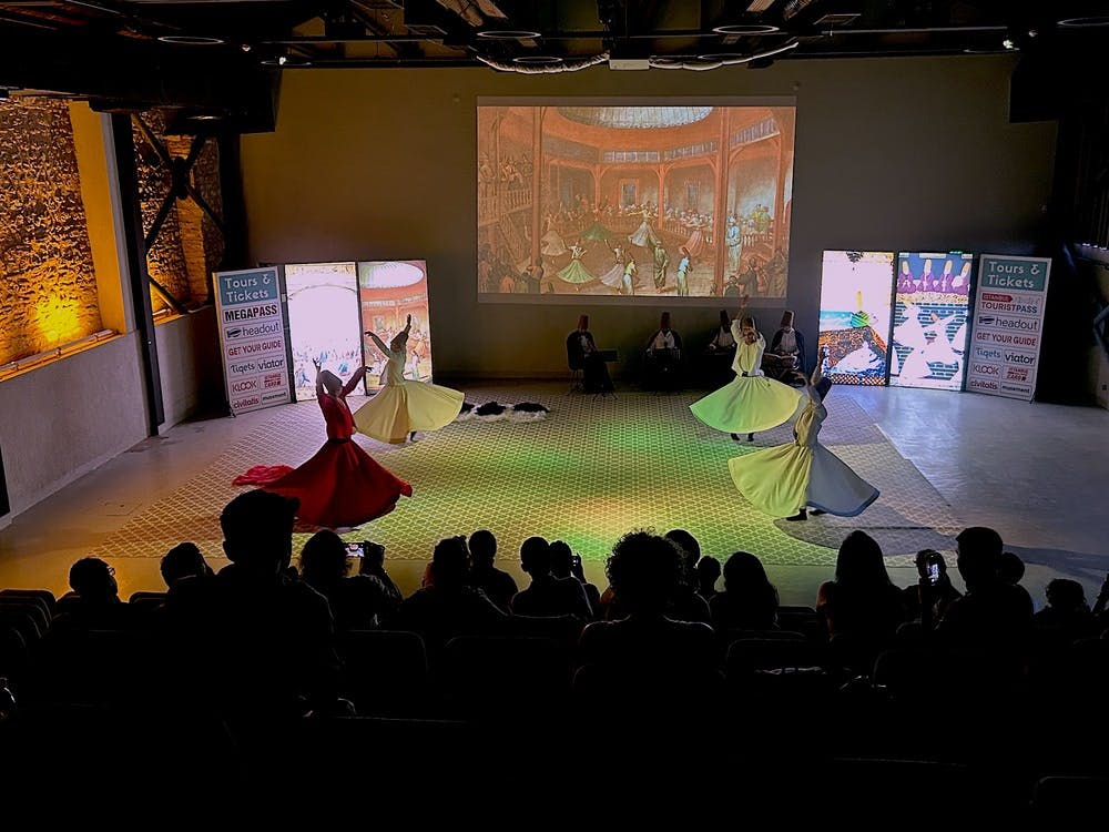 화려한 색상의 드레스를 입은 공연자들이 무대에서 춤을 추며 관객들의 시선을 사로잡습니다. 뒷벽에 역사적인 장면이 투영됩니다.