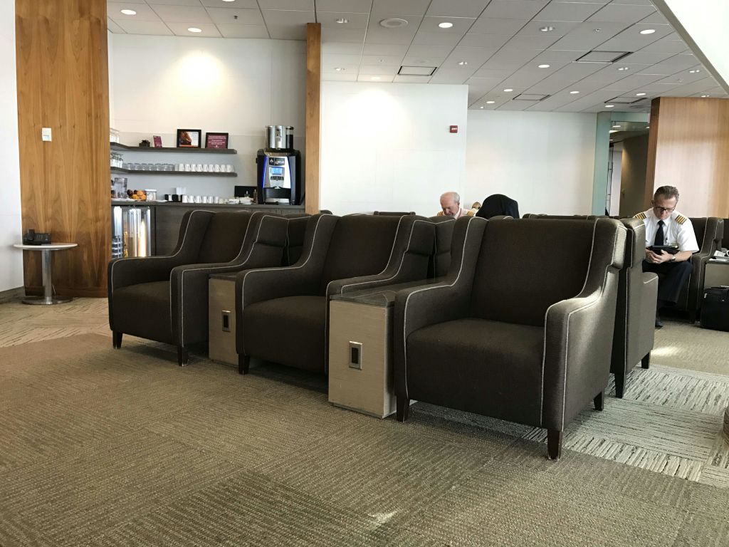 공항 라운지에 갈색 안락의자가 몇 개 있고, 두 명이 앉았으며, 한 명은 유니폼을 입고 있고, 한 명은 커피 스테이션을 배경으로 앉아 있습니다.
