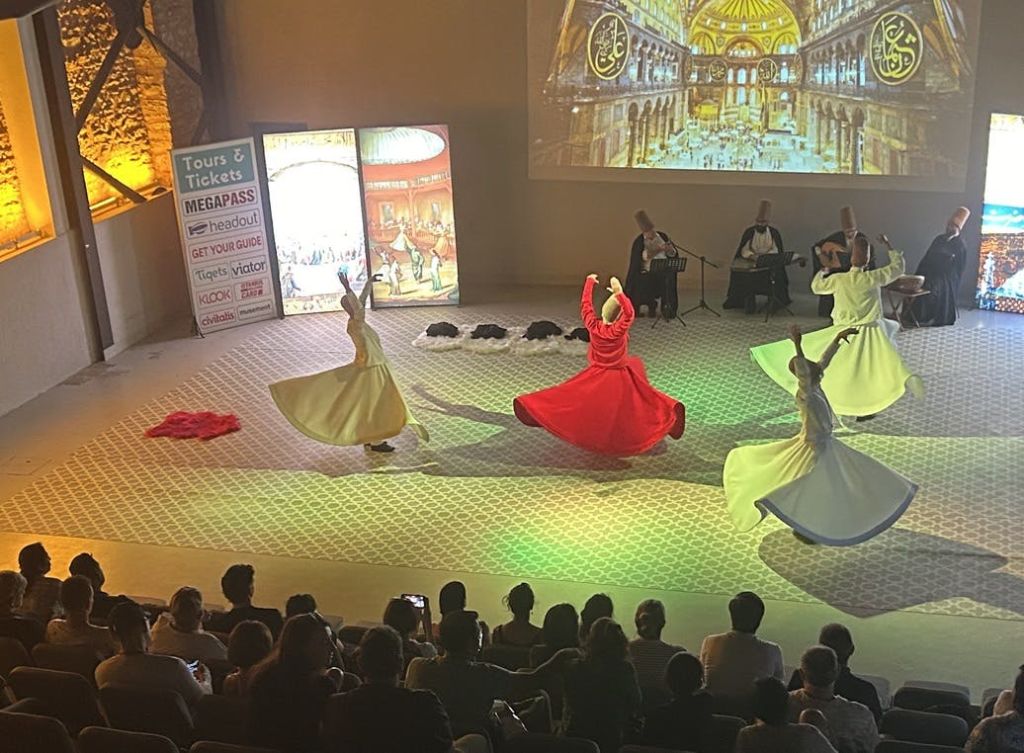 화려한 색상의 로브를 입은 댄서들이 무대에서 전통 스피닝 댄스를 선보입니다. 음악가를 배경으로 관객이 지켜보고 있습니다.