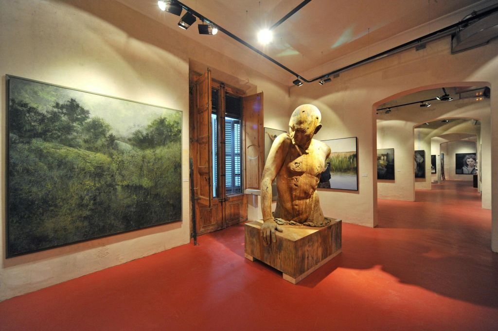 커다란 목조 인체 조각품, 벽에 그려진 풍경화, 빨간색 바닥에 설치된 트랙 조명이 특징인 갤러리입니다.