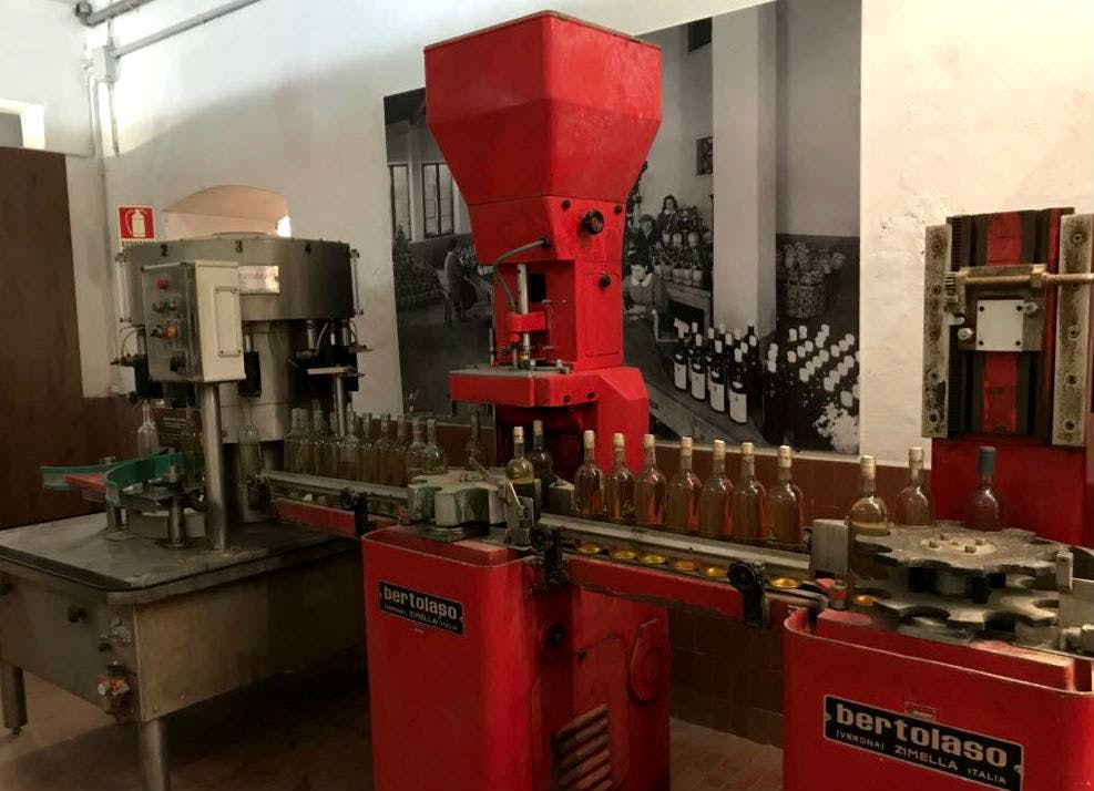 공장 설정에서 컨베이어 벨트에 병을 채우는 빨간색 Bertolaso 병입 기계. 배경은 쌓인 와인병을 보여줍니다.