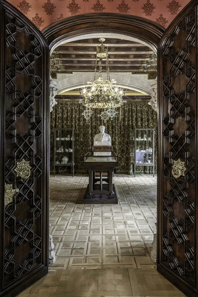 유리 디스플레이 케이스, 정교한 나무 문, 머리 위로 샹들리에가 있는 받침대에 가슴이 있는 화려한 객실입니다.