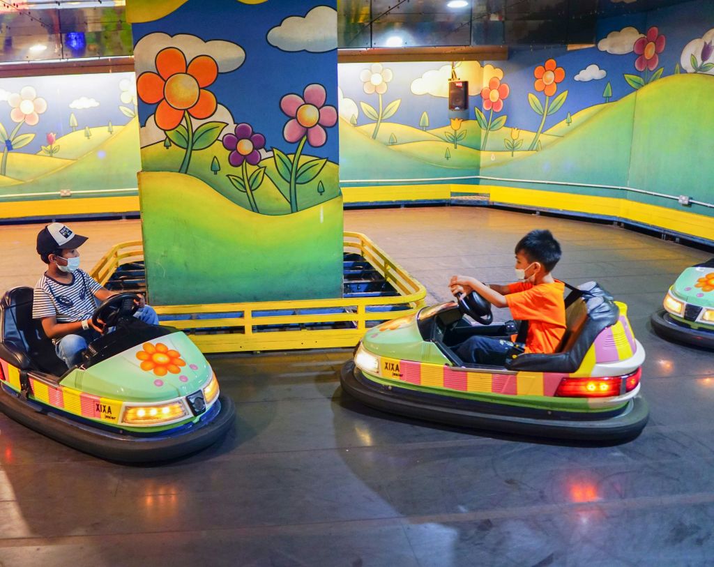 주황색 셔츠를 입은 어린이가 벽에 화려한 꽃 벽화가 걸린 실내 놀이 공간에서 범퍼 자동차를 타고 있습니다.
