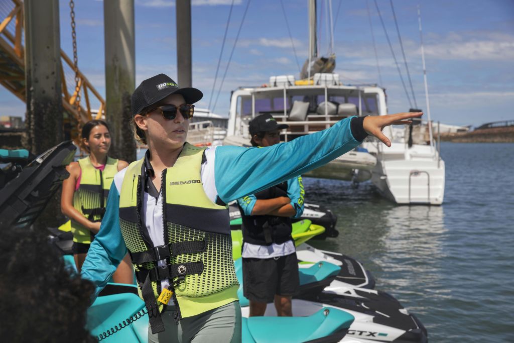 제트 스키 근처에서 구명 조끼를 입은 한 무리의 사람들이 다리와 보트를 배경으로 한 사람의 안내를 받습니다.
