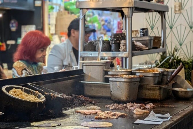 다양한 고기와 또띠아를 요리하는 그릴이 있는 음식 가판대입니다. 두 사람이 배경에 앉아 있고, 한 사람은 빨간 머리카락을 가지고 있습니다.