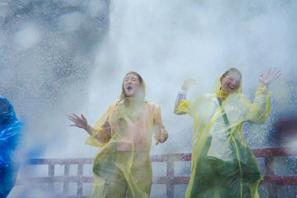 노란색 빗물 판초를 입은 두 사람이 나무 플랫폼에 서 있는 커다란 폭포에 흠뻑 젖고 있습니다.