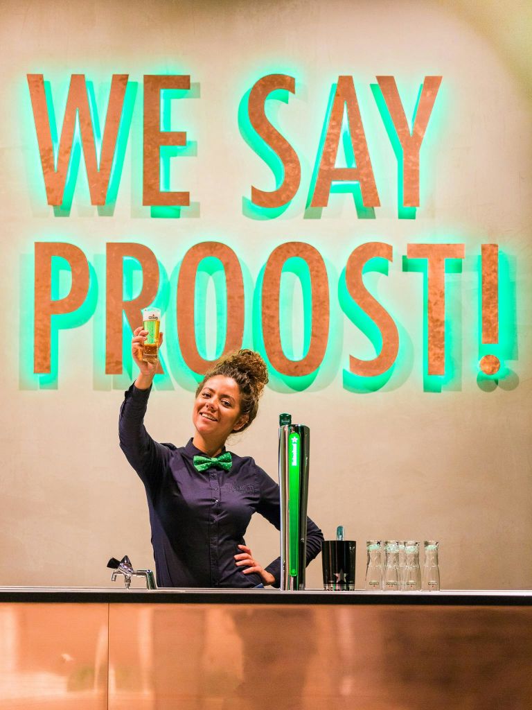 어두운 셔츠와 녹색 나비 넥타이를 매고 미소를 짓는 바텐더가 맥주 한 잔을 들고 있습니다. "우리는 PROOST를 말한다!" 그녀의 뒤쪽 벽에 불이 들어왔다.