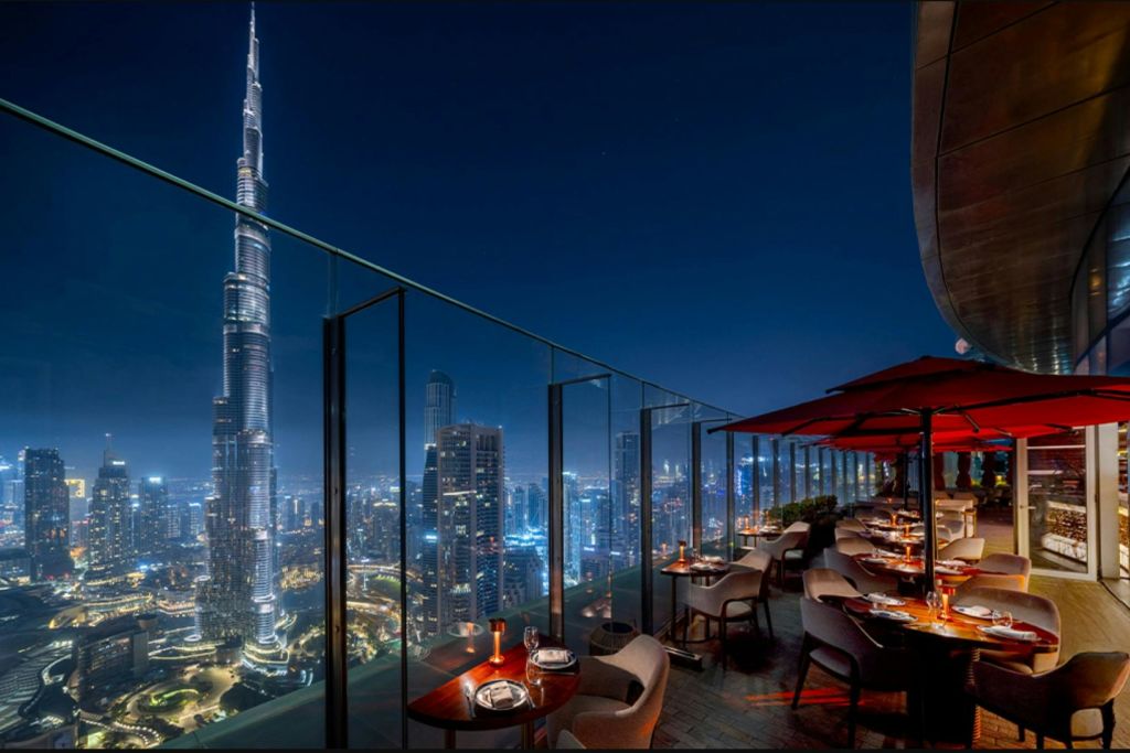 도시 전망을 감상할 수 있는 야외 옥상 레스토랑으로 부르즈 할리파와 붉은 우산이 놓인 조명 테이블이 있습니다.