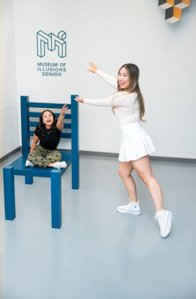 두 사람이 커다란 착시 의자를 들고 포즈를 취하고 있는데, 한 사람은 그 위에 앉아 있고, 다른 한 사람은 덴버 환상 박물관 간판을 가리키고 있습니다.