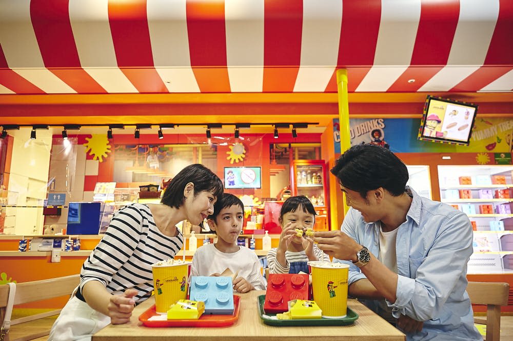 4인 가족이 빨간색과 흰색 줄무늬 장식이 있는 활기찬 레고 테마 레스토랑에서 식사를 하고 있습니다. 레고 모양의 요리가 있습니다.
