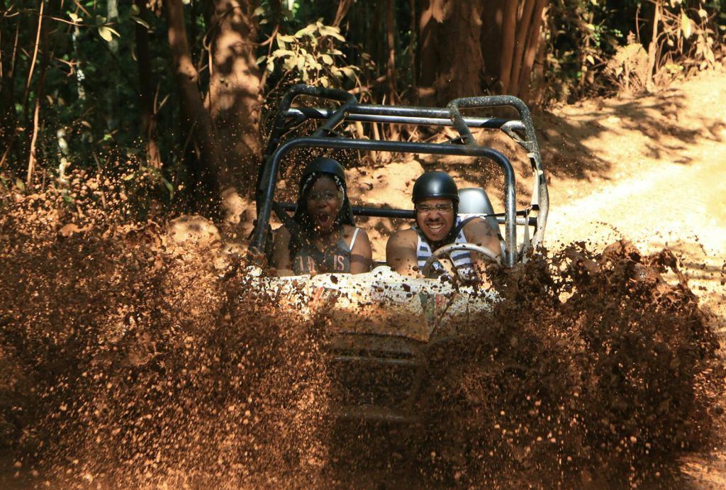 헬멧을 쓴 두 사람이 모래 언덕 버기를 몰고 진흙 지형을 지나 숲속에서 진흙을 튀깁니다.