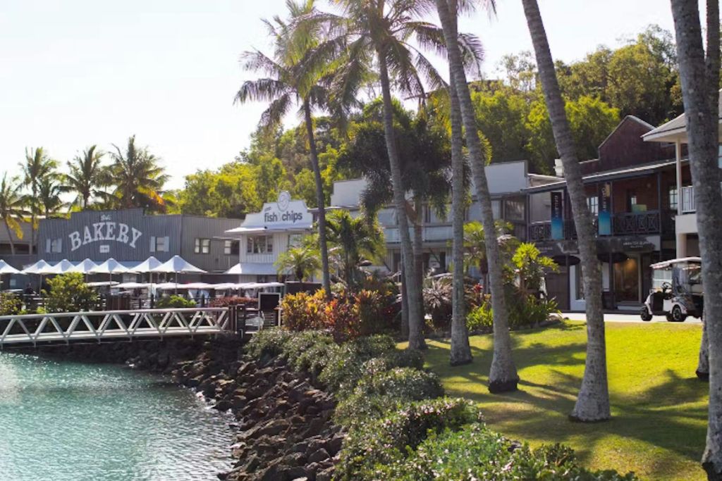 야자수, 피시앤칩스 가게, 텐트 아래 야외 좌석, 물가 산책로가 있는 해안 풍경.