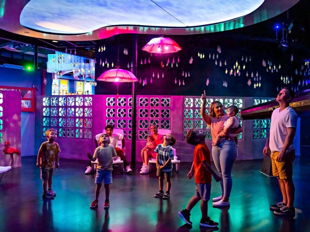 화려한 조명, 분홍색 우산, 장식용 벽 패널이 있는 밝은 방에서 어린이와 성인이 고개를 들고 있습니다.