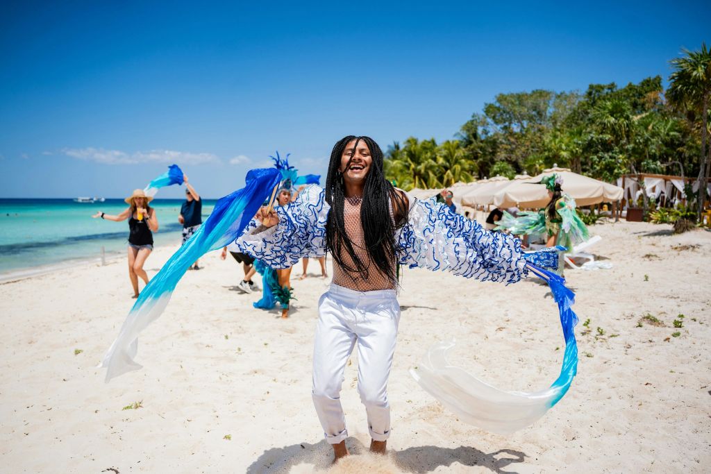 파란색과 녹색 액세서리로 다른 사람들에게 둘러싸인 흰색 바지와 장식 상의를 입고 해변에서 즐겁게 춤을 추는 사람.