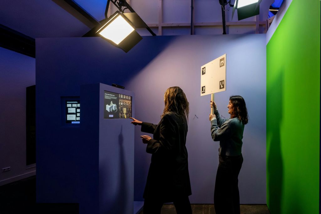 두 사람이 파란색 전시 부스와 상호 작용합니다. 하나는 스크린을 가리키고 다른 하나는 마커 보드를 높이 들고 있습니다.