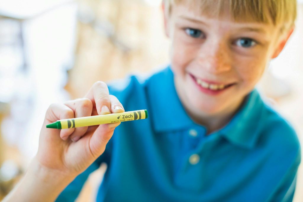 '자크' 라고 적힌 노란색 연두색 크레용을 카메라 가까이에 둔 파란색 셔츠를 입고 미소를 짓고 있는 아이.