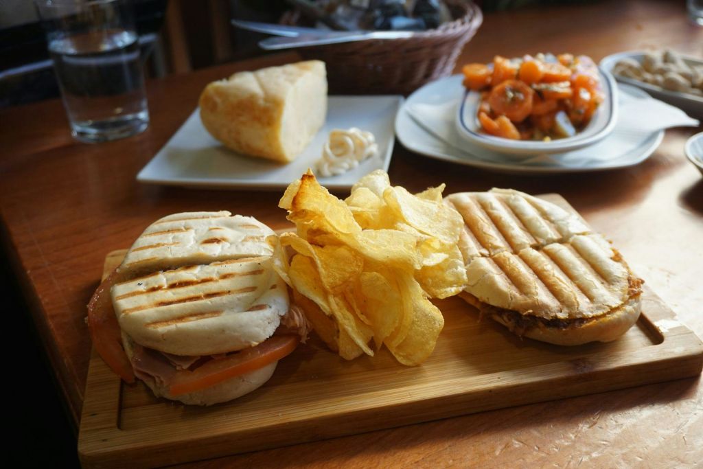 두 개의 파니니 샌드위치, 감자 칩, 샐러드가 담긴 접시, 나무 테이블 위에 놓인 치즈 블록으로 구성된 식사.