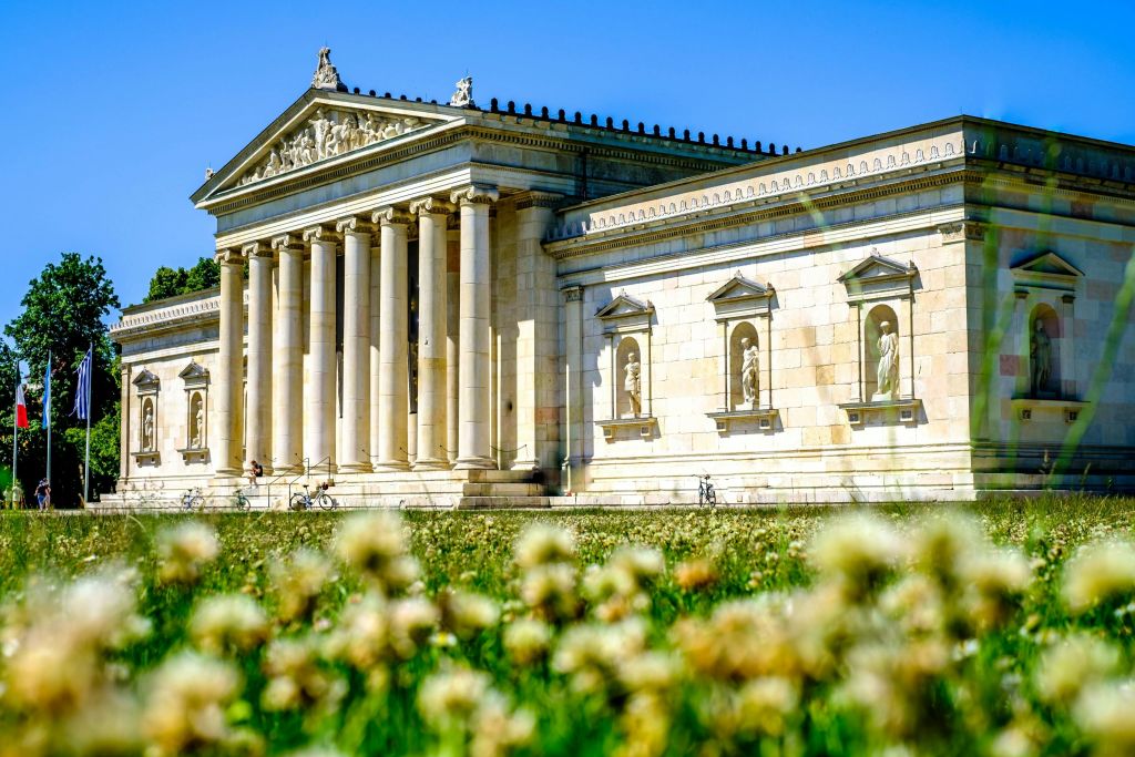 맑고 푸른 하늘 아래 기둥과 벽 틈새에 있는 동상이 있는 신고전주의 양식의 건물. 전경의 꽃과 녹지.