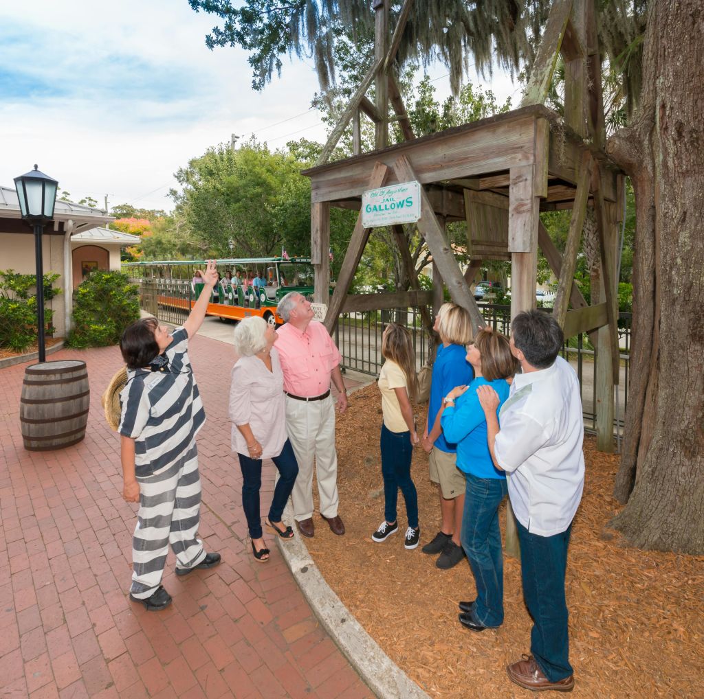 죄수로 옷을 입은 한 사람이 역사적인 야외 환경에서 여섯 명의 사람들에게 "올드 세인트 어거스틴 갤로우스" 표지판을 지적합니다.