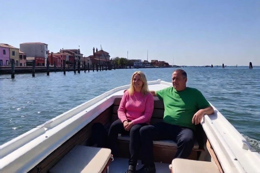 맑은 하늘 아래 배경으로 건물과 나무가 보이는 잔잔한 물 속 보트에 앉아 있는 두 사람.