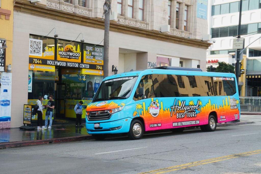 다채로운 색상의 할리우드 투어 버스가 도시 거리의 방문자 센터 근처에 주차되어 있습니다. 건물 근처에 간판과 관광객이 있습니다.