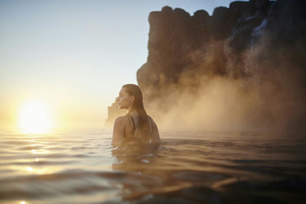 안개가 솟구치고 바위가 많은 절벽을 배경으로 해돋이에 온천을 즐기는 여성.